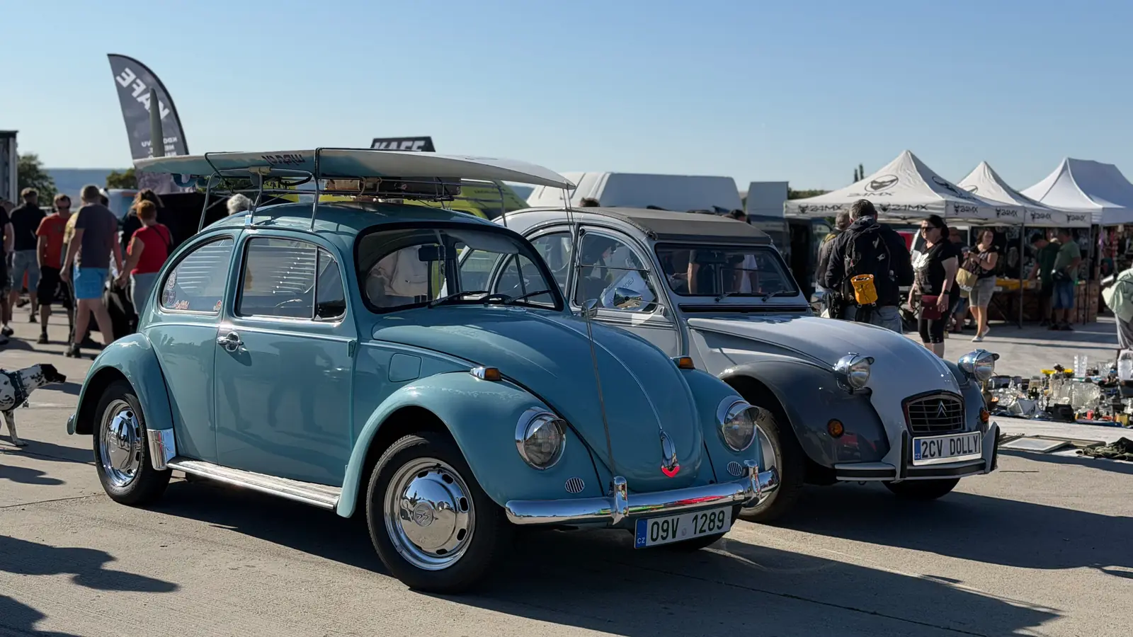 Modrý VW Brouk zepředu se surfem na střeše, vedle stříbrný Citroën 2CV